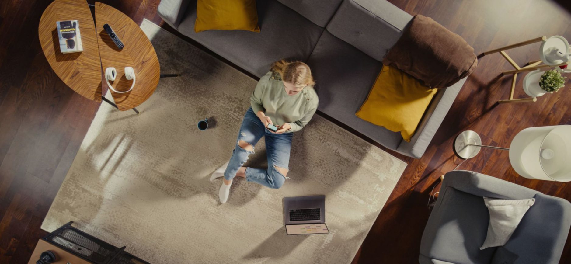 Wohnzimmer-Szene von oben aufgenommen. Auf einem großen hellen Teppich sitzt eine Person auf dem Boden, angelehnt an ein dunkelgraues Sofa. Die Person hat blonde Haare, trägt einen hellgrünen Pullover, blaue zerissene Jeans und weiße Socken. In den Händen hält sie ein Smartphone, der Blick ist darauf gerichtet, die Beine sind locker angewinkelt. Links vor ihr steht ein aufgeklappter Laptop auf dem Teppich.