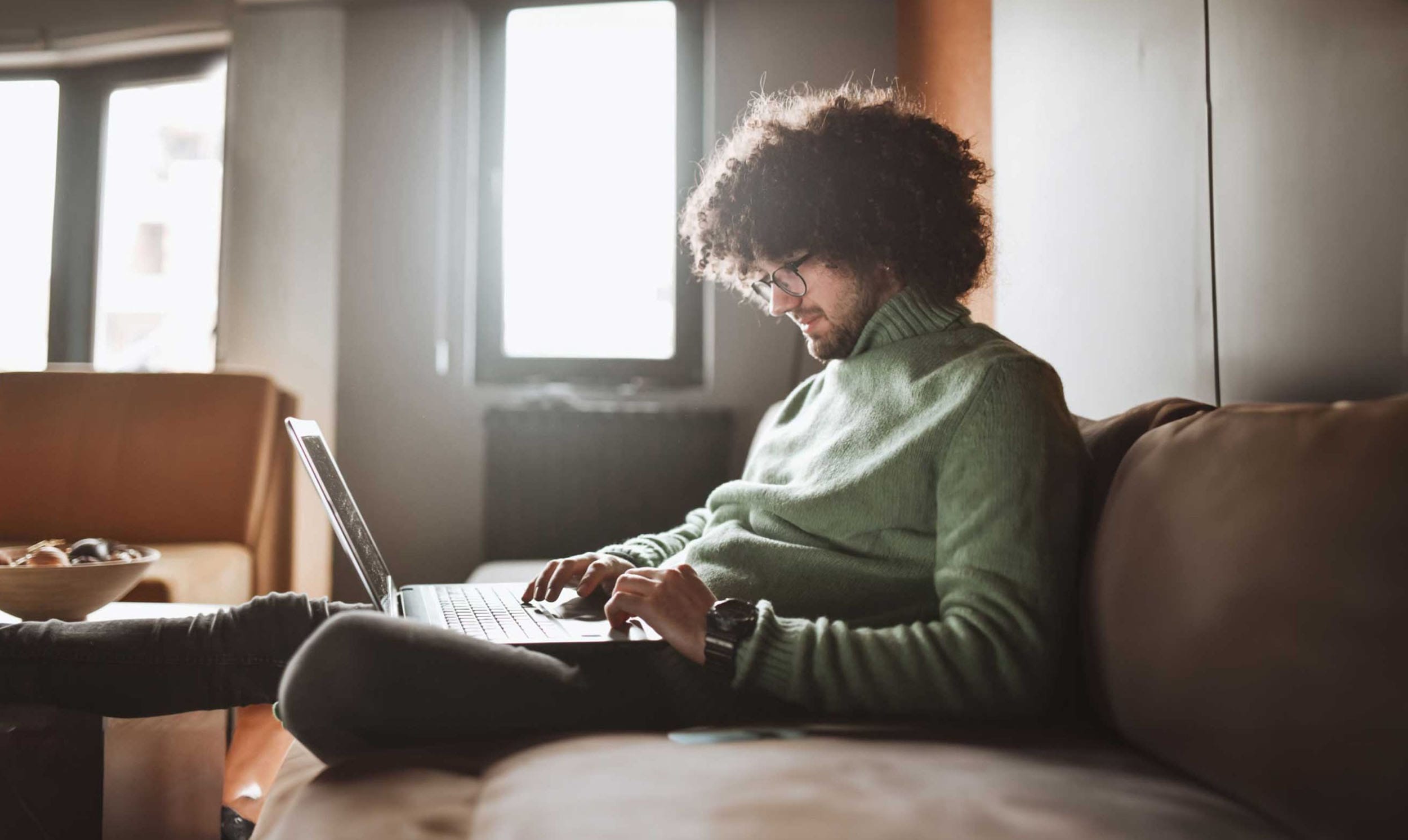 Ein junger Mann mit lockigem, dunklen Haar und hellem Teint sitzt entspannt auf einem Sofa und arbeitet an einem Laptop.
