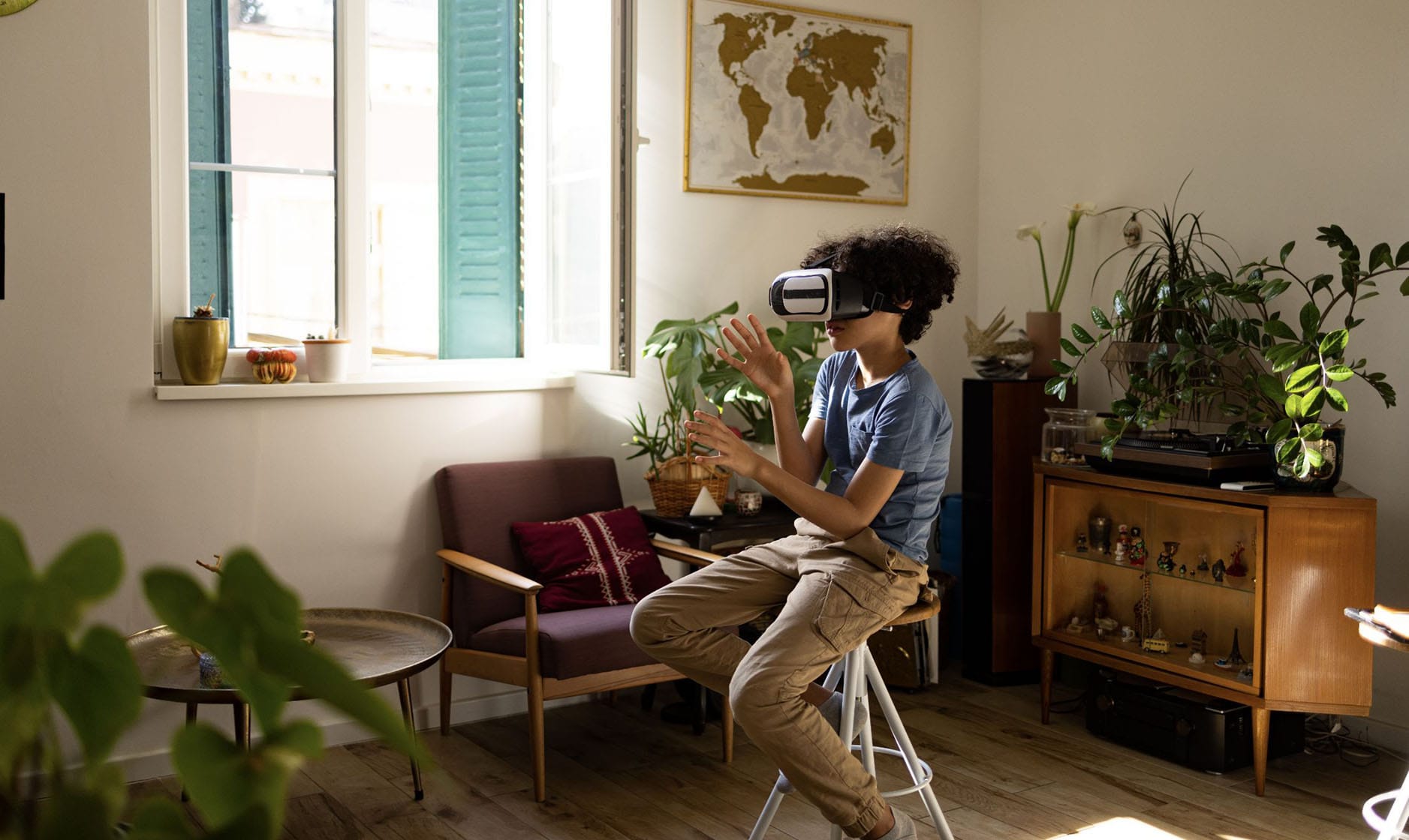 Eine Person mit Virtual-Reality-Brille sitzt in einem Wohnzimmer. Die Person ist schlank, hat mittleren Teint und dichtes, dunkles, lockiges Haar. Sie sitzt auf einem weißen Klapphocker, trägt ein blaues kurzärmliges Hemd und beigefarbene Cargohosen. Die Hände sind in der Luft erhoben, die Finger gespreizt, als würde etwas Unsichtbares berührt oder gesteuert.