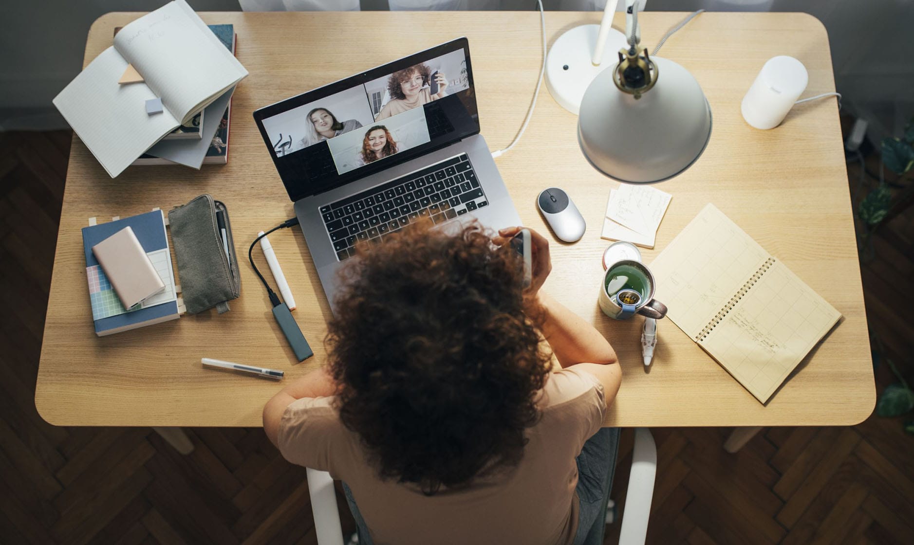 Schreibtisch mit aufgeklapptem Laptop von oben fotografiert. Auf dem Laptop läuft ein Videoanruf mit drei Personen, deren Gesichter in separaten Fenstern zu sehen sind. Vor dem Laptop liegen eine Computermaus, ein Smartphone und ein kleiner Adapter. Auf dem Schreibtisch liegen mehrere Bücher, Notizblöcke, Zettel und Stifte.