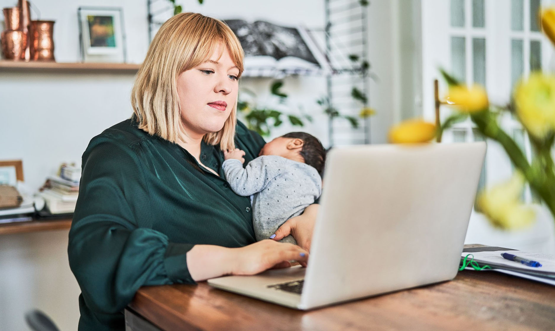 Eine blonde Frau mit hellbeiger Haut sitzt an einem Holztisch und hält ein kleines, schlafendes Baby im Arm. Vor der Frau steht ein aufgeklappter, hellfarbener Laptop auf dem Tisch.