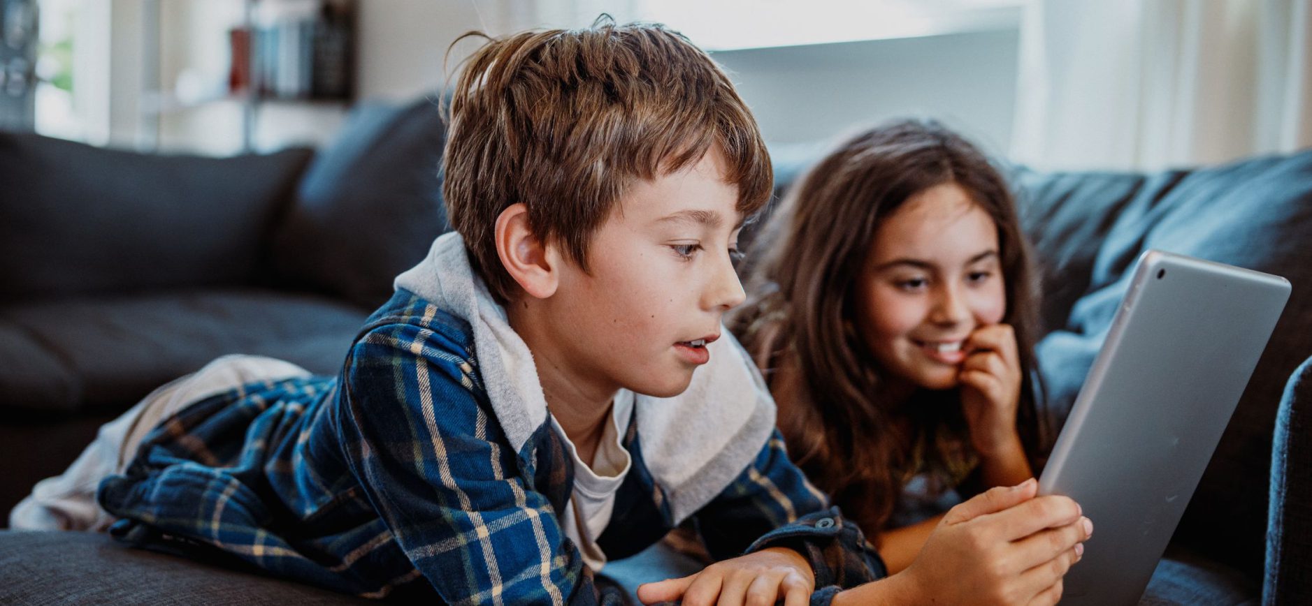 Zwei Kinder auf einem Sofa, die gemeinsam auf ein Tablet schauen. Im Vordergrund liegt ein Junge mit hellbraunen Haaren seitlich auf einem Sofa, den Kopf leicht nach unten geneigt. Sein Gesichtsausdruck wirkt konzentriert. Neben ihm, etwas darunter, liegt ein Mädchen mit langen, dunkelbraunen Haaren. Sie stützt den Kopf mit einer Hand ab und lächelt, während sie auf den Bildschirm schaut.