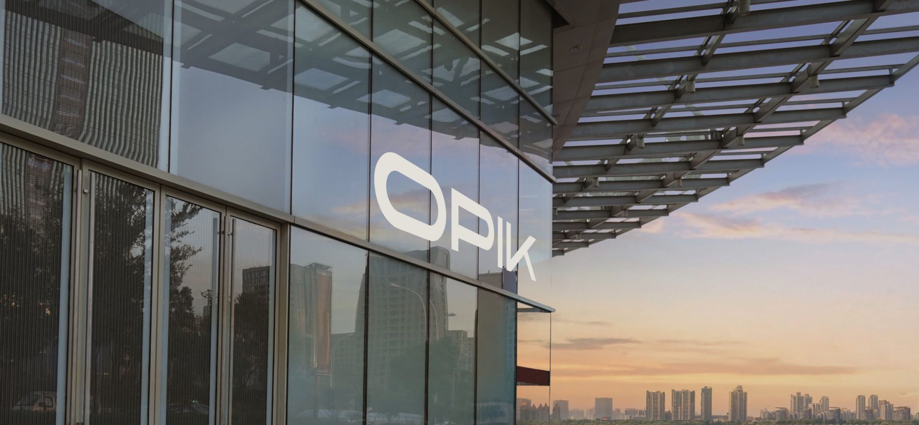 Glasfassade eines modernen Hochhauses mit großem, weißen Schriftzug "OPIK". Die Fassade besteht aus großen Glasflächen und Metallstreben, die schräg nach oben laufen und ein Raster bilden. Im Glas spiegeln sich andere Hochhäuser und Bäume. Es ist ein Himmel mit leicht bewölktem Sonnenuntergang im Hintergrund zu sehen und eine Skyline mit mehreren schlanken Hochhäusern.